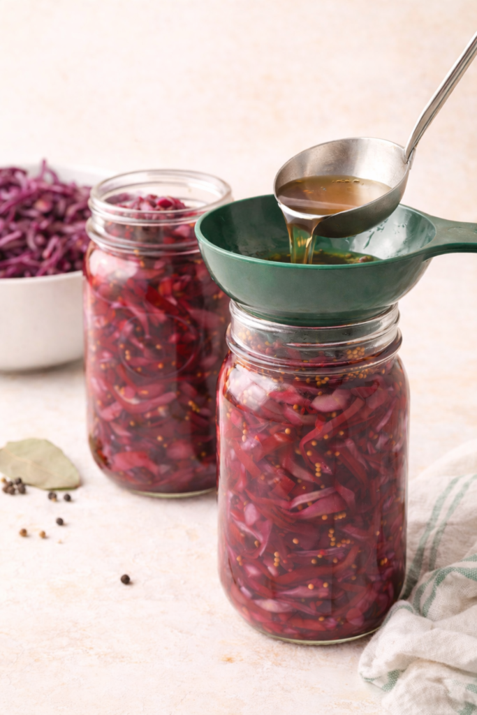 Pouring warm spiced vinegar brine into mason jars filled with shredded red cabbage using a funnel for water bath canning.