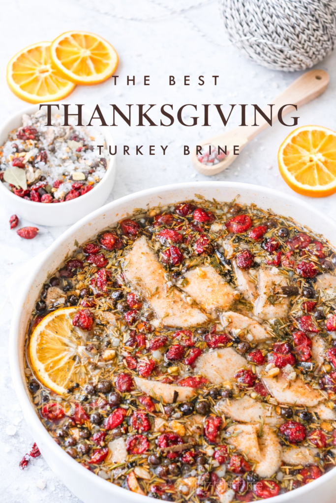 Raw turkey soaking in a flavorful Thanksgiving turkey brine with cranberries, herbs, citrus, and spices in a white bowl on a marble countertop