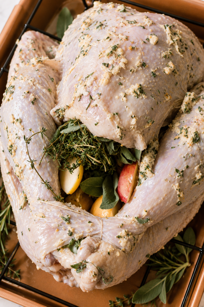 Raw Thanksgiving turkey coated with herb butter and stuffed with fresh herbs, garlic, apples, and citrus before roasting.
