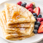 Easy homemade crepes drizzled with maple syrup and dusted with powdered sugar on a white platter with fresh strawberries, raspberries, and blueberries.