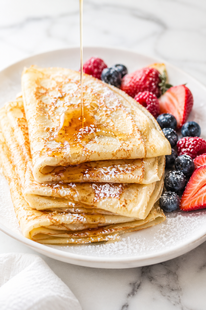 Easy homemade crepes drizzled with maple syrup and dusted with powdered sugar on a white platter with fresh strawberries, raspberries, and blueberries.