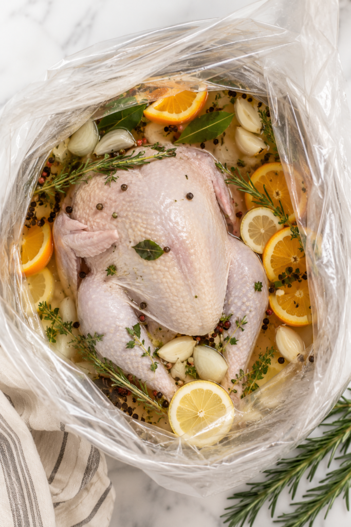 Whole turkey soaking in a citrus herb brine with oranges, lemons, garlic, rosemary, thyme, and peppercorns inside a brining bag on a marble countertop.