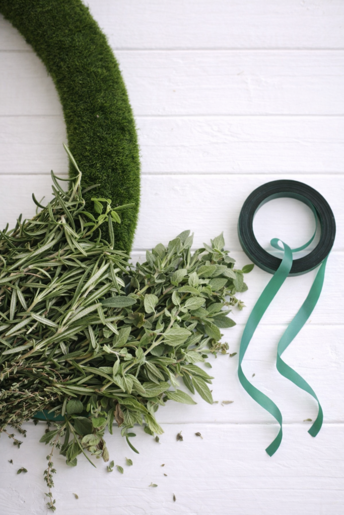 Fresh rosemary and oregano attached to a moss wreath form with floral tape on a white background, showing a DIY herb wreath in progress.