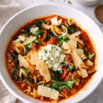 Bowl of lasagna soup topped with herbed ricotta, shaved parmesan, fresh basil, Italian sausage, egg noodles, and tomato broth on a light marble countertop.