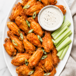 Crispy baked buffalo chicken wings coated in homemade buffalo sauce and served on a white oval platter with celery sticks and ranch dressing on a white marble countertop.