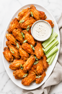 Crispy baked buffalo chicken wings coated in homemade buffalo sauce and served on a white oval platter with celery sticks and ranch dressing on a white marble countertop.