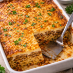 Greek pastitsio baked in a white casserole dish with a slice removed, showing layers of pasta, spiced meat sauce, and creamy béchamel
