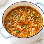 Hearty lentil and brown rice soup simmering in a light enamel cast iron pot with vegetables and herbs, bright and cozy healthy soup.