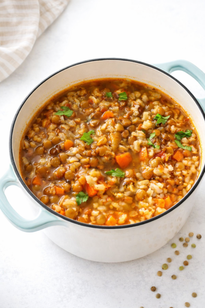 Hearty lentil and brown rice soup simmering in a light enamel cast iron pot with vegetables and herbs, bright and cozy healthy soup.