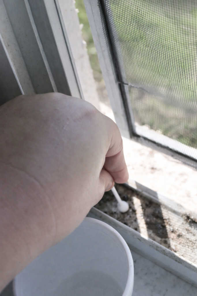 Hand using a cotton swab to clean dirt and grime from a window track during deep cleaning in natural light