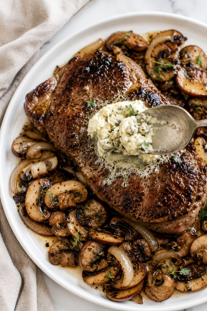 A spoon adds creamy garlic Gorgonzola butter onto a hot pan-seared steak with sautéed mushrooms and onions on a white plate.