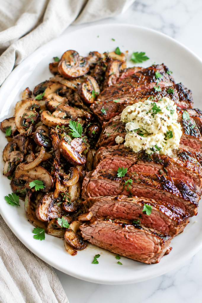 Juicy pan-seared steak topped with creamy garlic Gorgonzola butter and served with sautéed mushrooms and onions on a white plate.