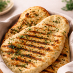 Freshly baked cilantro and garlic naan stacked in a basket with grill marks and herbs, soft and golden with a rustic homemade look.