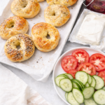 Freshly baked homemade bagels on a parchment-lined baking sheet with cream cheese, sliced tomatoes, cucumbers, and pickled red onions.