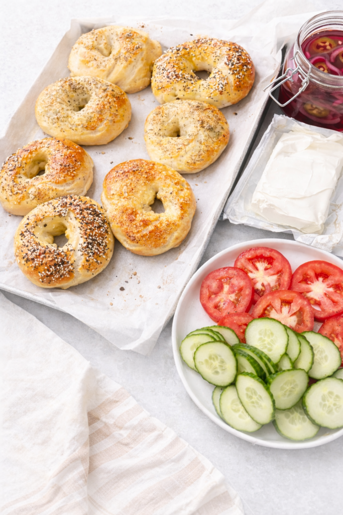 Freshly baked homemade bagels on a parchment-lined baking sheet with cream cheese, sliced tomatoes, cucumbers, and pickled red onions.