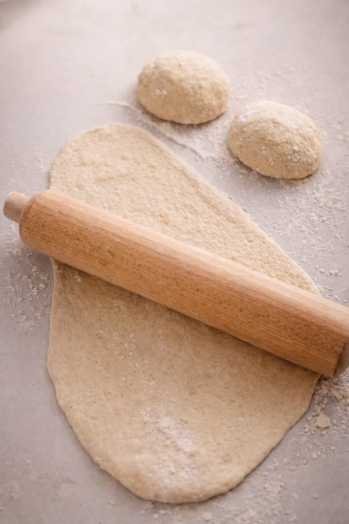 Rolling out naan dough with a wooden rolling pin on a lightly floured surface, showing the shaping process for homemade flatbread.