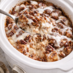 Creamy cinnamon roll bread pudding scooped from a white slow cooker with pecans and vanilla glaze