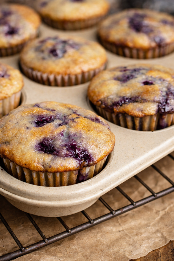 Blueberry jam muffins baked in a beige muffin pan, with golden tops and swirls of blueberry jam, cooling on a wire rack