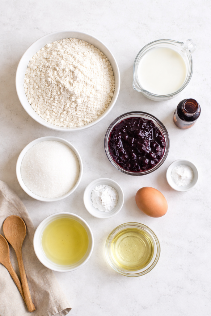 Flat lay of ingredients for blueberry jam muffins arranged on a light marble countertop with bowls of flour, sugar, milk, oil, egg, vanilla, and blueberry jam