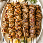 Grilled Greek chicken and pork souvlaki skewers served on a white platter with homemade garlicky tzatziki, fresh oregano, lemon wedges, and charred juicy meat on a marble countertop.