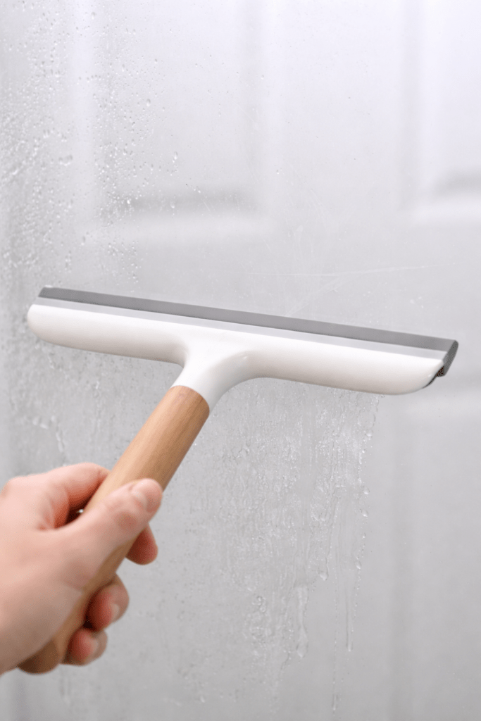 Modern white and wood handle squeegee removing water and soap scum from glass shower door in a bright clean bathroom.