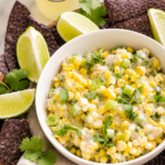 Creamy spicy ranch corn salsa in a white bowl with blue corn tortilla chips and fresh lime wedges on a marble countertop.