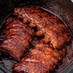 Three racks of fall-off-the-bone BBQ ribs cooking in a slow cooker with glossy sauce