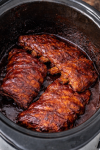 Three racks of fall-off-the-bone BBQ ribs cooking in a slow cooker with glossy sauce