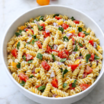 Garlic lovers pasta salad in a large white bowl on a marble counter with fresh tomatoes, herbs, and creamy dressing