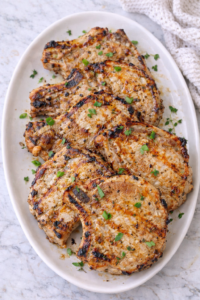 Grilled maple Dijon pork chops arranged on a large white platter with fresh herbs.