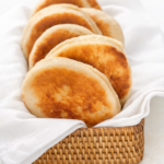 Golden homemade English muffins in a white cloth-lined wicker basket on a light neutral background