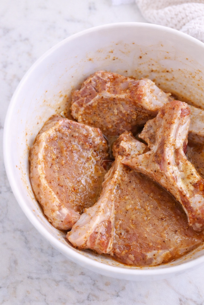 Pork chops marinating in maple Dijon glaze in a white bowl on a marble countertop.