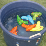 Colorful squirt guns floating in a blue bucket filled with water on green grass, set up as an easy summer squirt gun refill station for kids.