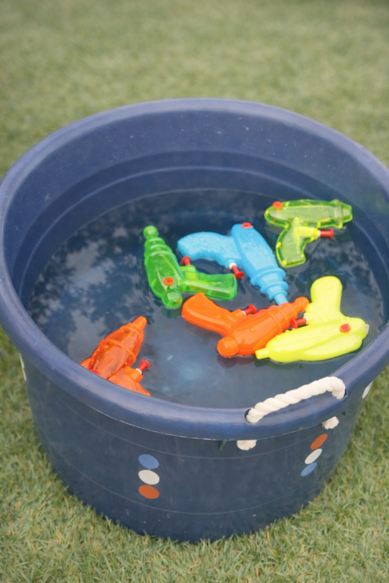Colorful squirt guns floating in a blue bucket filled with water on green grass, set up as an easy summer squirt gun refill station for kids.