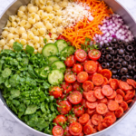 Deli style pasta salad ingredients in a bowl before mixing including pasta, vegetables, pepperoni, olives, and dressing on a marble counter