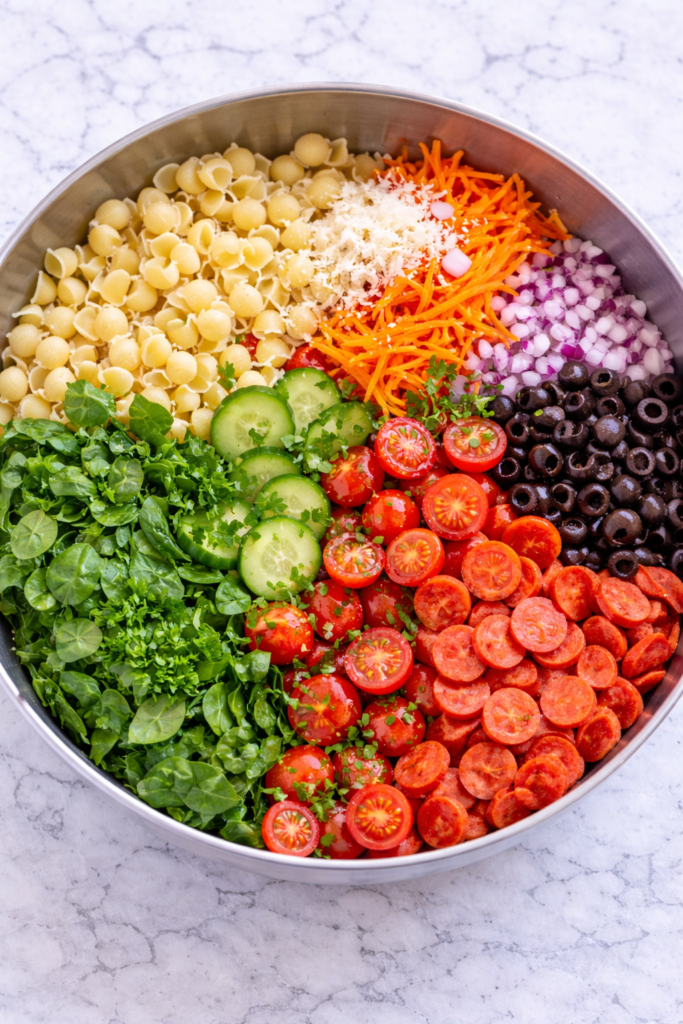 Deli style pasta salad ingredients in a bowl before mixing including pasta, vegetables, pepperoni, olives, and dressing on a marble counter