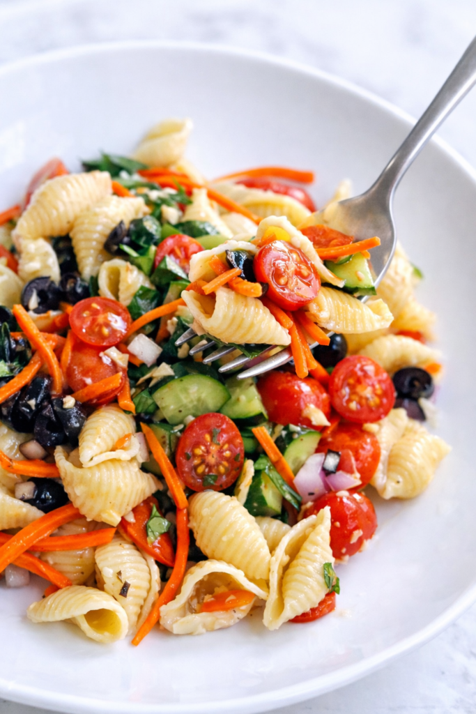 Deli style pasta salad with shells, vegetables, and dressing being lifted on a fork from a white bowl