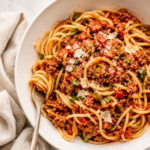 Large white bowl of spaghetti topped with hearty ground turkey and sausage meat sauce on a white marble countertop with a neutral linen cloth.