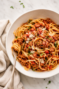 Large white bowl of spaghetti topped with hearty ground turkey and sausage meat sauce on a white marble countertop with a neutral linen cloth.