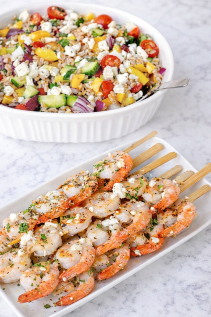 Greek grilled shrimp and farro salad with feta, cucumber, tomatoes, and lemon on a marble countertop, served with herb grilled shrimp skewers