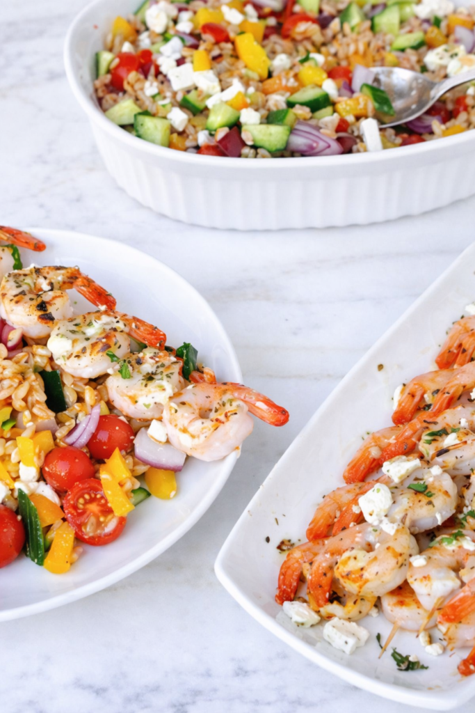 Greek shrimp farro salad served in white bowls with grilled shrimp, feta, cucumber, tomatoes, and red onion on a marble countertop
