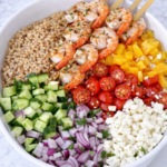 Greek shrimp farro salad ingredients in a bowl before mixing with farro, cucumber, tomatoes, red onion, feta, and grilled shrimp on a marble countertop