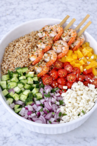 Greek shrimp farro salad ingredients in a bowl before mixing with farro, cucumber, tomatoes, red onion, feta, and grilled shrimp on a marble countertop