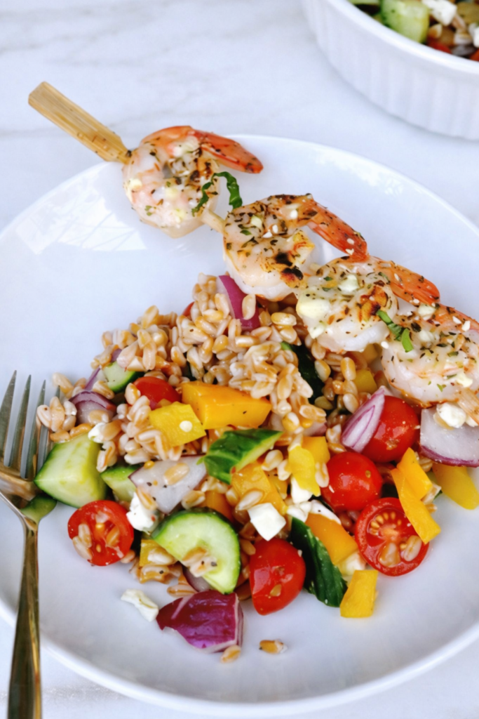 Greek shrimp farro salad served on a white plate with grilled shrimp skewer, fresh vegetables, and feta on a marble countertop