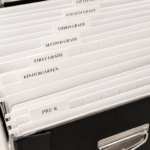 White hanging file folders labeled by grade inside a black school paperwork storage box for organizing children's school papers and keepsakes.