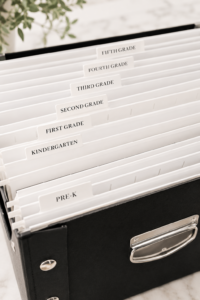 White hanging file folders labeled by grade inside a black school paperwork storage box for organizing children's school papers and keepsakes.