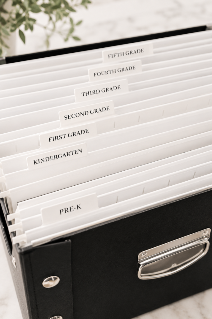White hanging file folders labeled by grade inside a black school paperwork storage box for organizing children's school papers and keepsakes.