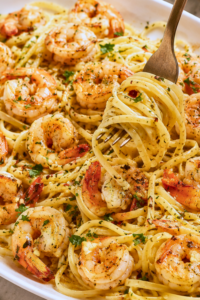 Fork twirling linguine topped with garlic butter shrimp scampi on a large white platter with fresh parsley, Parmesan, lemon, and red pepper flakes.