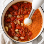 A white immersion blender pureeing roasted tomatoes, onions, garlic, and herbs in a white enamel Dutch oven on a white marble countertop.