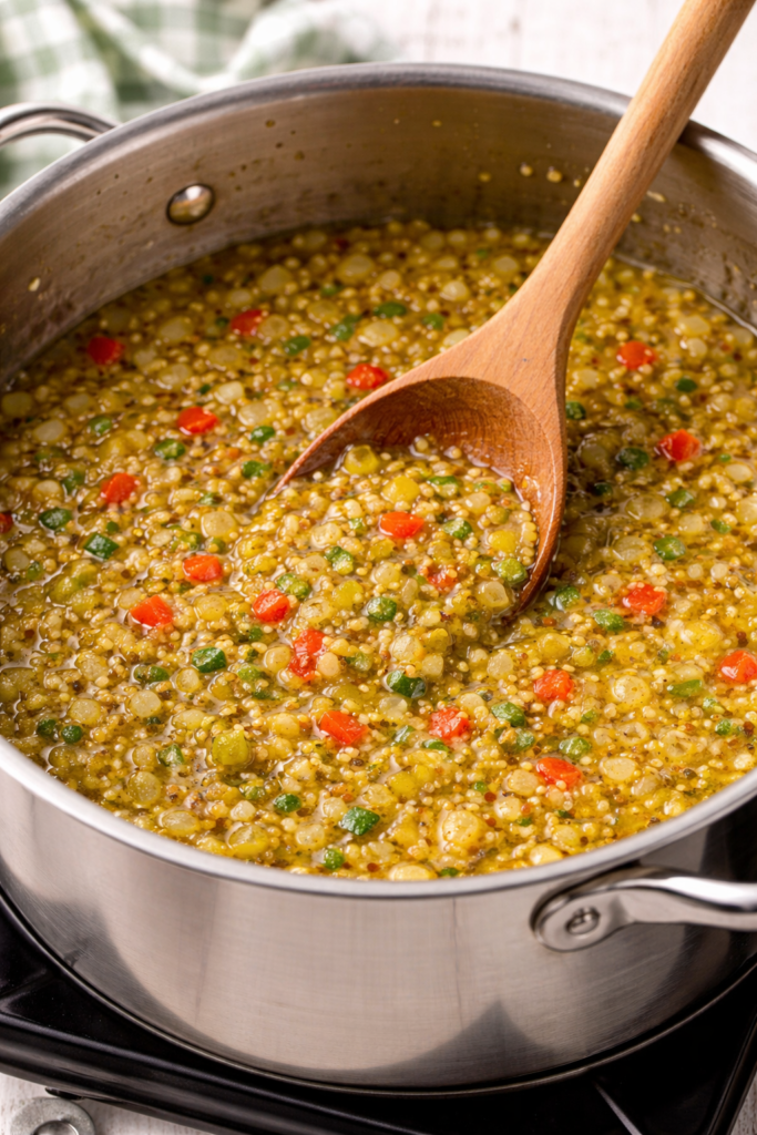 Homemade zucchini relish simmering in a stainless steel pot on the stove with a wooden spoon.